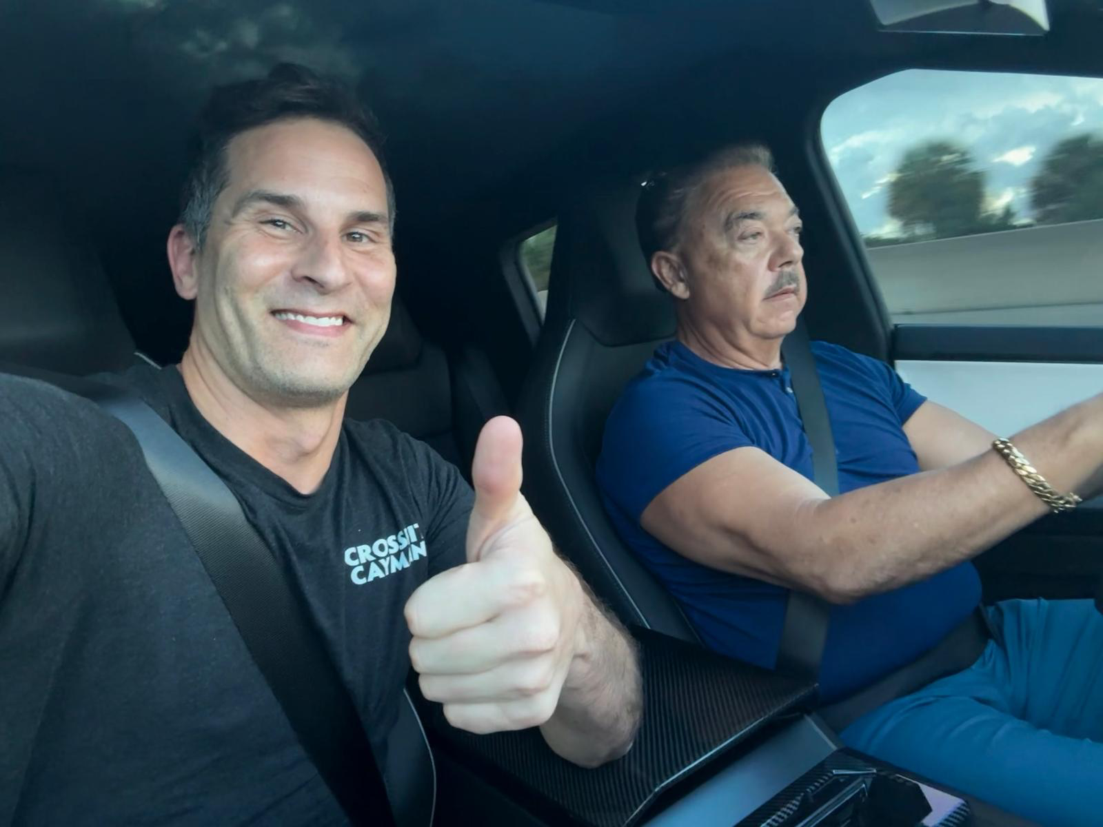 Passenger in a CrossFit Cayman tee giving a thumbs up; driver in a blue polo focused on the road