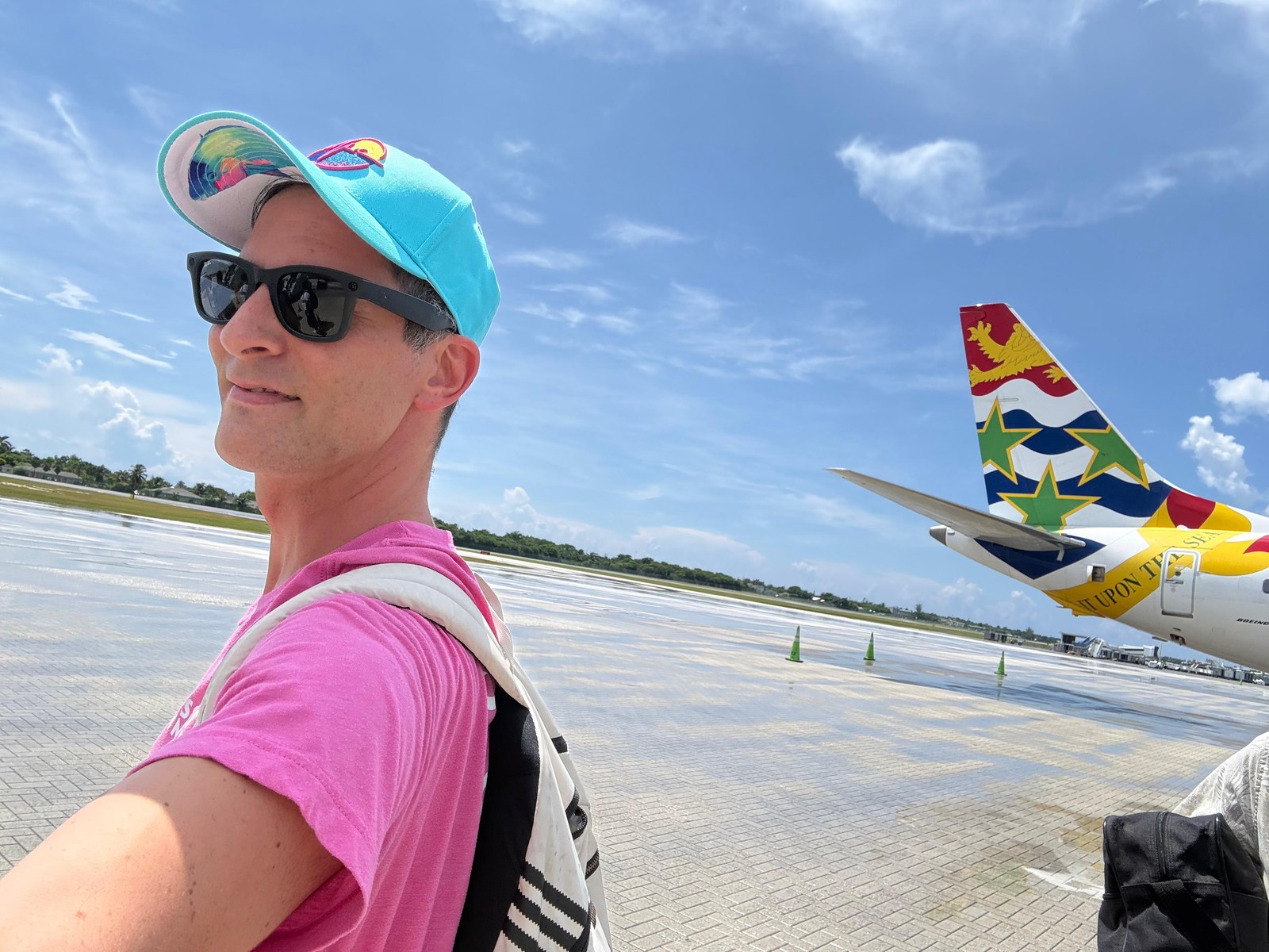 Person in a pink tee and turquoise cap with a white backpack on the tarmac, Cayman Airways tail behind them